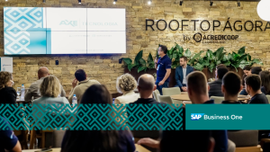 Apresentação da Áxe Tecnologia em evento no Rooftop Ágora by Acredicoop, com público assistindo à palestra sobre soluções SAP Business One em um ambiente corporativo moderno e acolhedor.